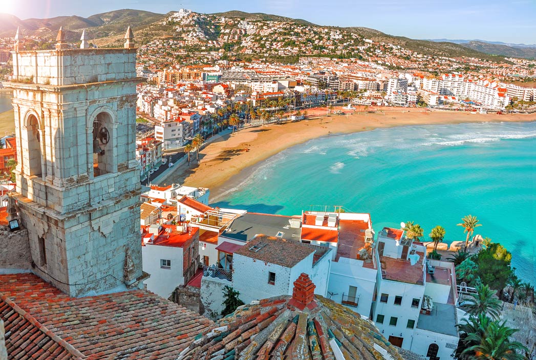 A picturesque coastal city with white buildings lines a sandy beach, where turquoise water meets the shore. In the foreground, there's a historic stone church tower that rises above the seaside promenade. Rolling hills stretch into the distance, providing a scenic backdrop to the vibrant waterfront scene.