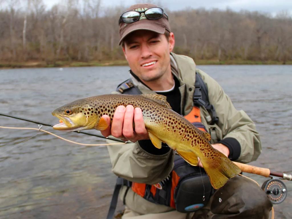 A photo featuring a fly fishing angler standing in the river and posing while holding a Brown Trout in one hand above the water