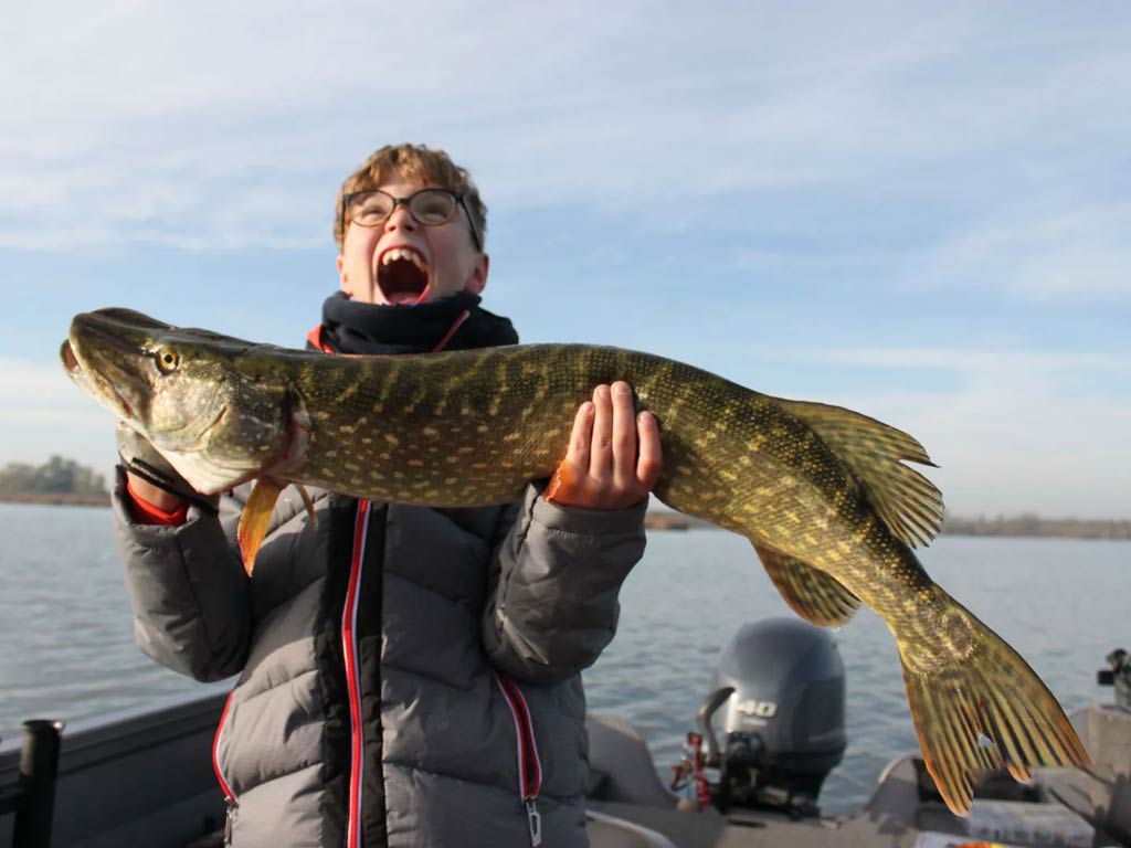 A photo featuring a happy kid while standing on a charter boat and posing with a big Pike he caught while fishing in the Netherlands