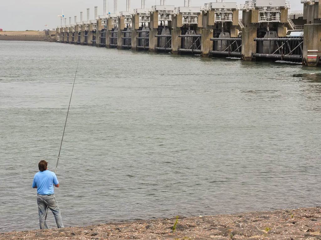 A photo featuring and angler while standing on the shore and fishing next to the Great Dike of Oosterschelde