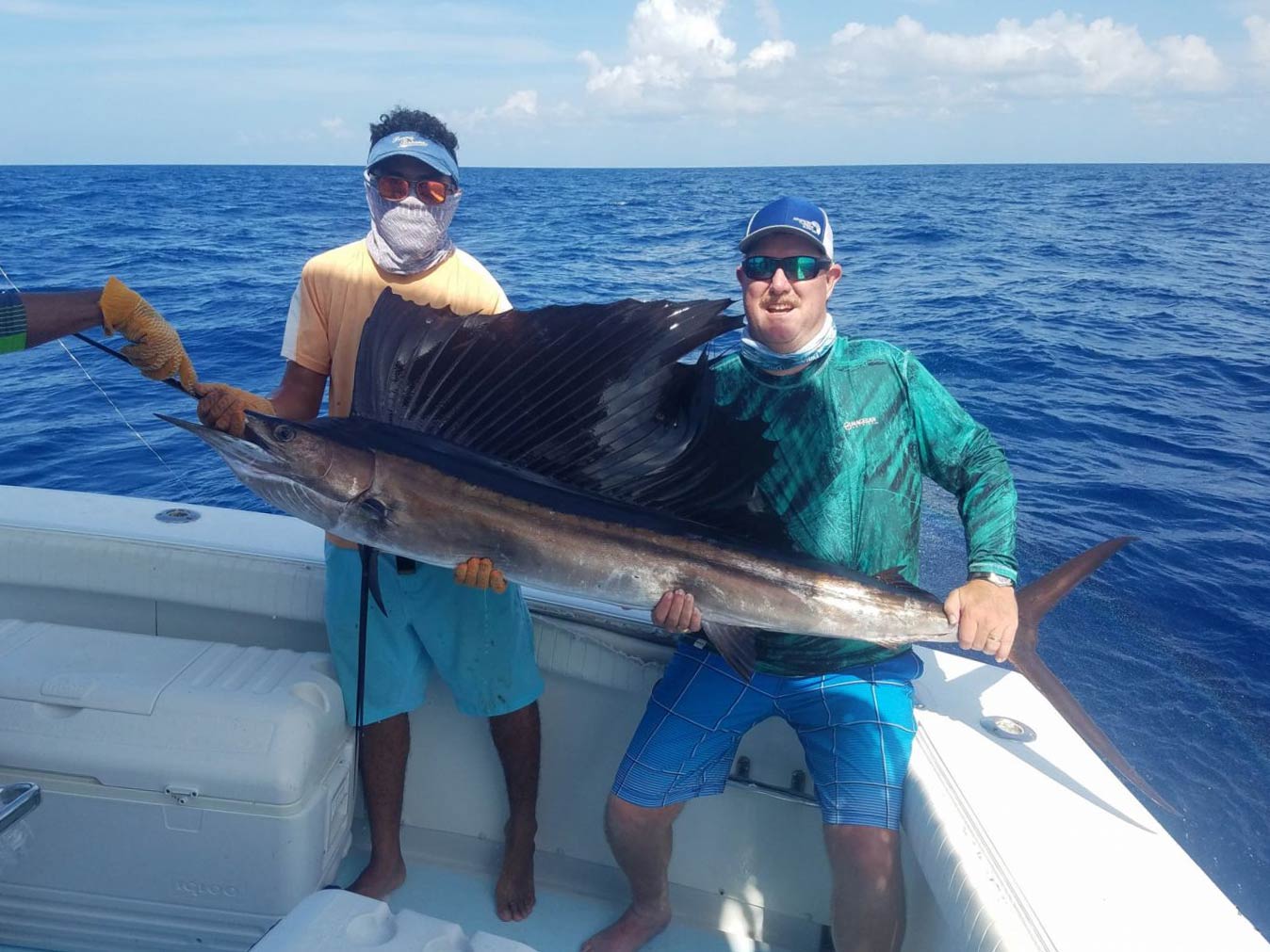 Two anglers standing on a boat holding a massive Sailfish, with water in the background