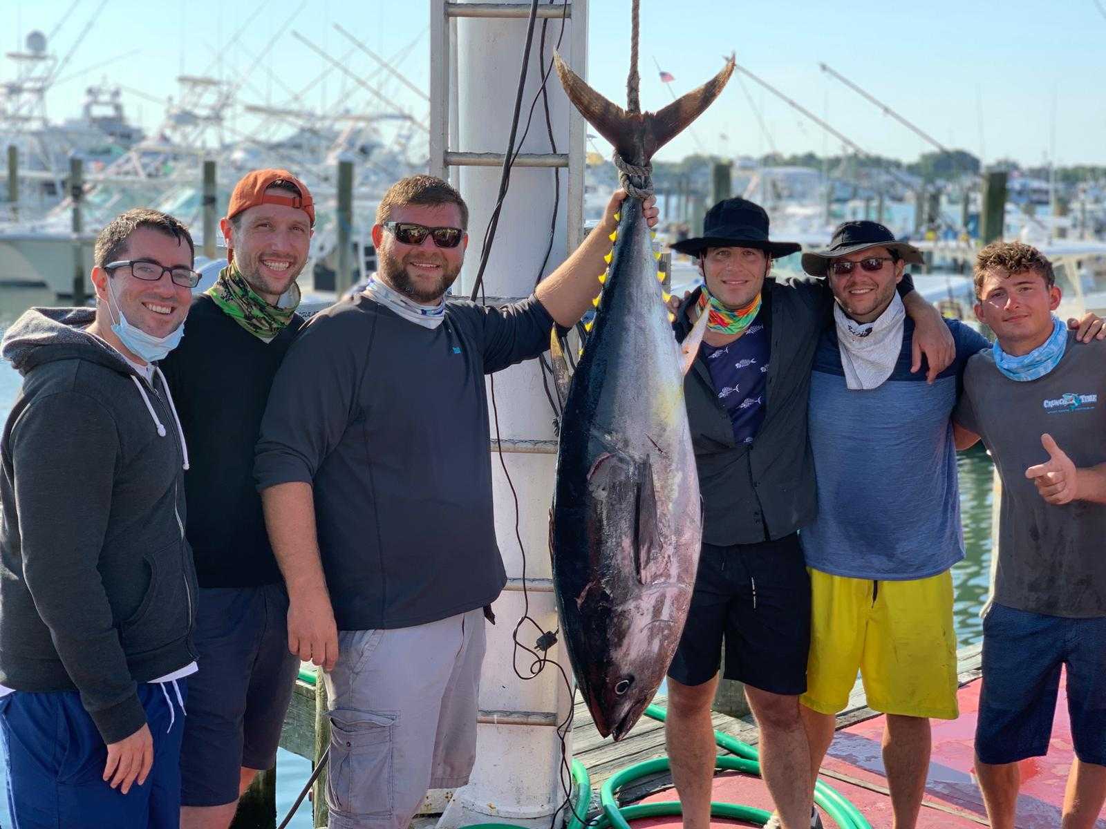 A group of friends with charter crew standing on a dock holding a big Bluefin Tuna hanged by the tail in between them
