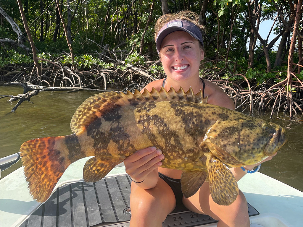 A woman sat on an inshore fishing boat in front of some mangrove trees, holding a Gag Grouper on a sunny day.