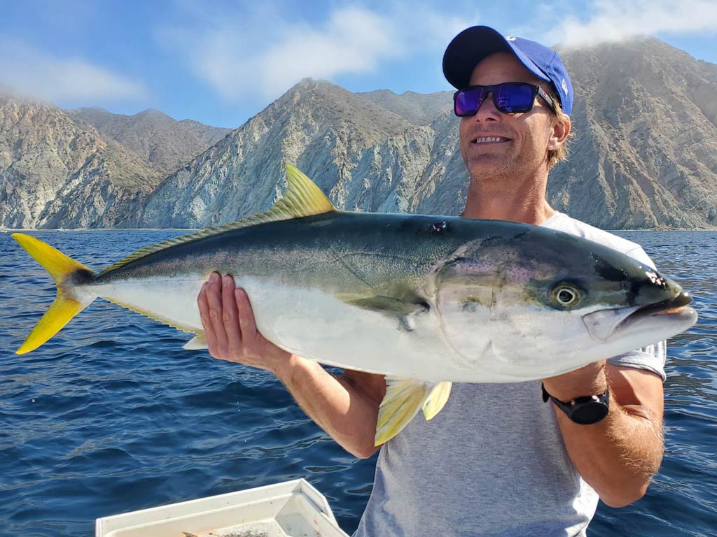 A photo of an angler proudly posing while holding a trophy Yellowtail Amberjack, with the Catalina shoreline in the background on a bright and sunny day
