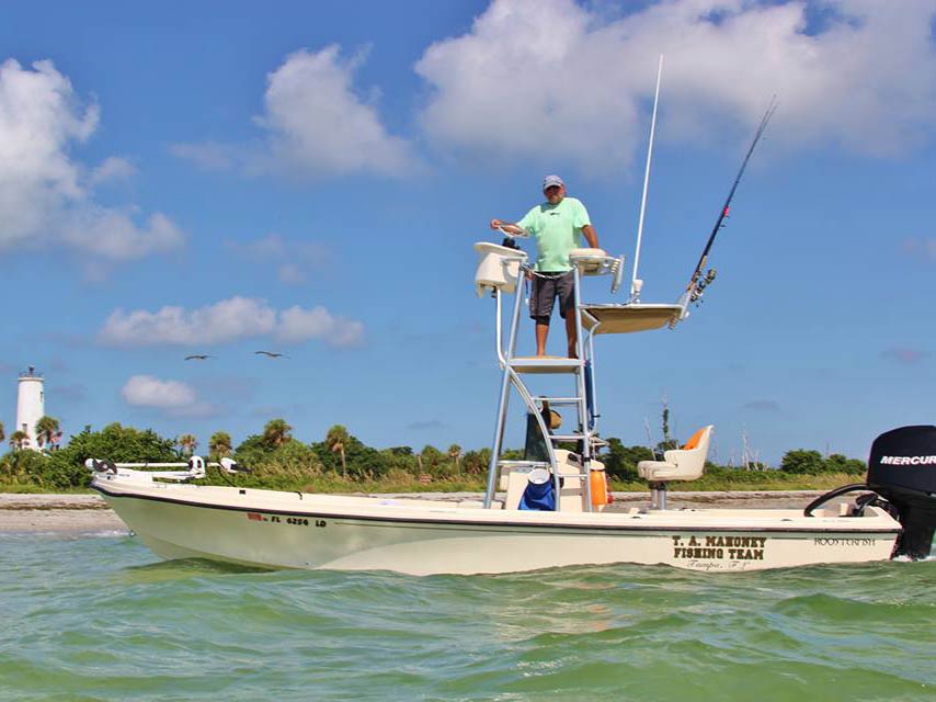 A captain stands high on his platform on a boat in the inshore waters, with a beach in the background and a lighthouse on the left of the image