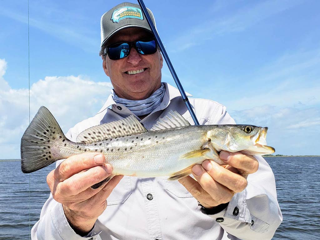 A photo of a happy and smiling angler wearing a cap and a pair of sunglasses while standing on a charter boat and proudly showing off Speckled Trout 