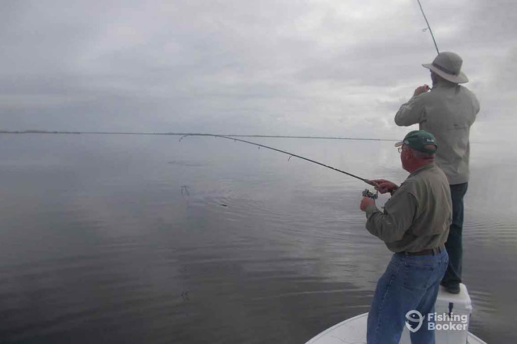 Two anglers standing on a small fishing charter in the Mosquito Lagoon, casting their lines into the calm waters on a cloudy day