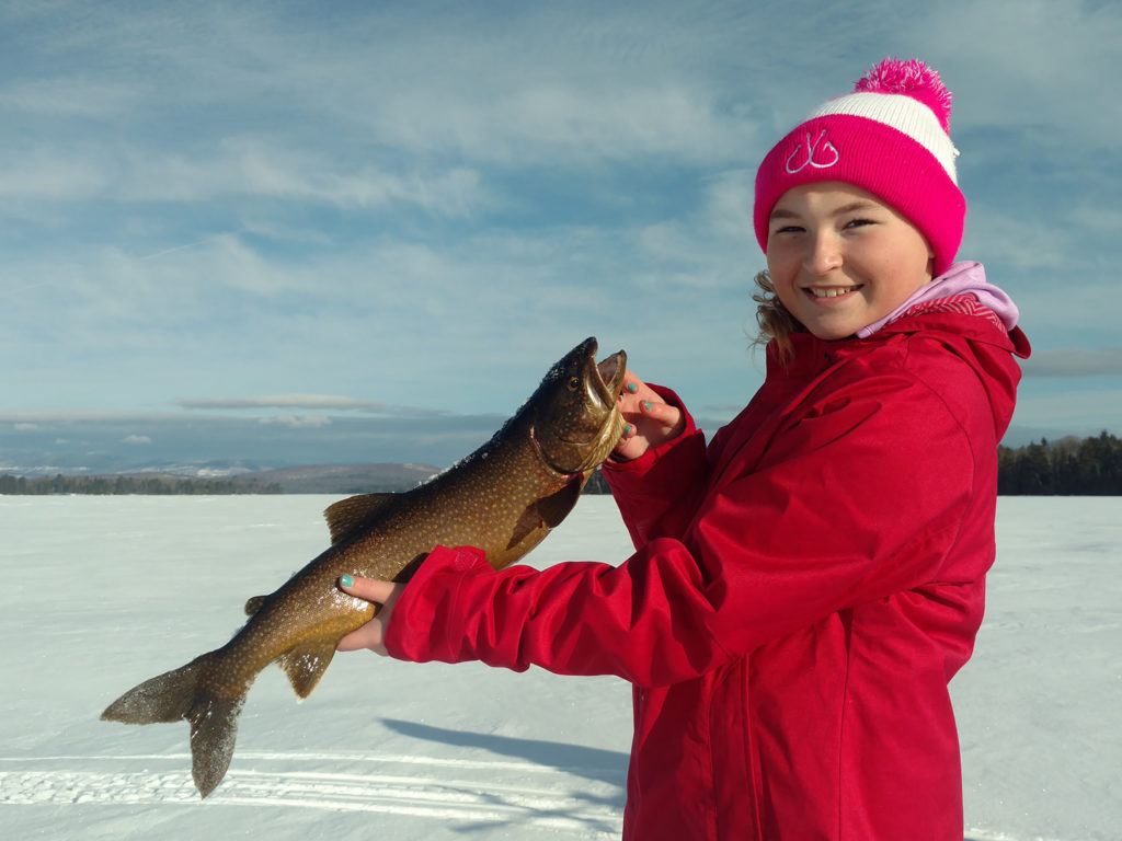 A young girl holding a Lake Trout on an ice fishing trip on a frozen lake.