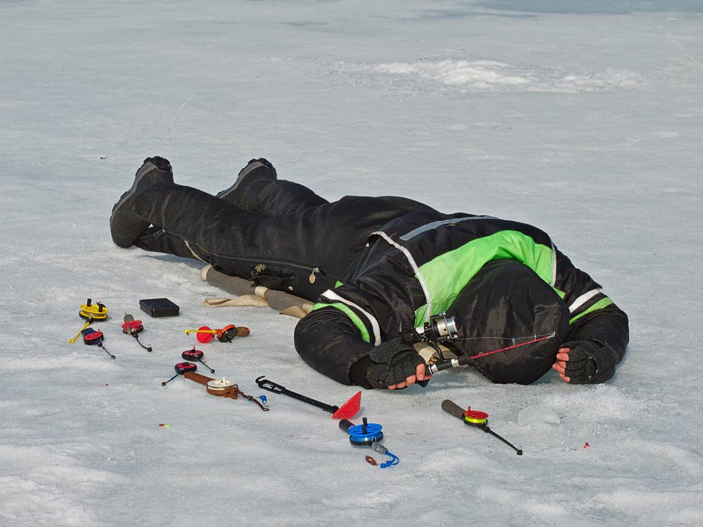 An ice angler lying on the frozen lake and looking down at a hole in the ice
