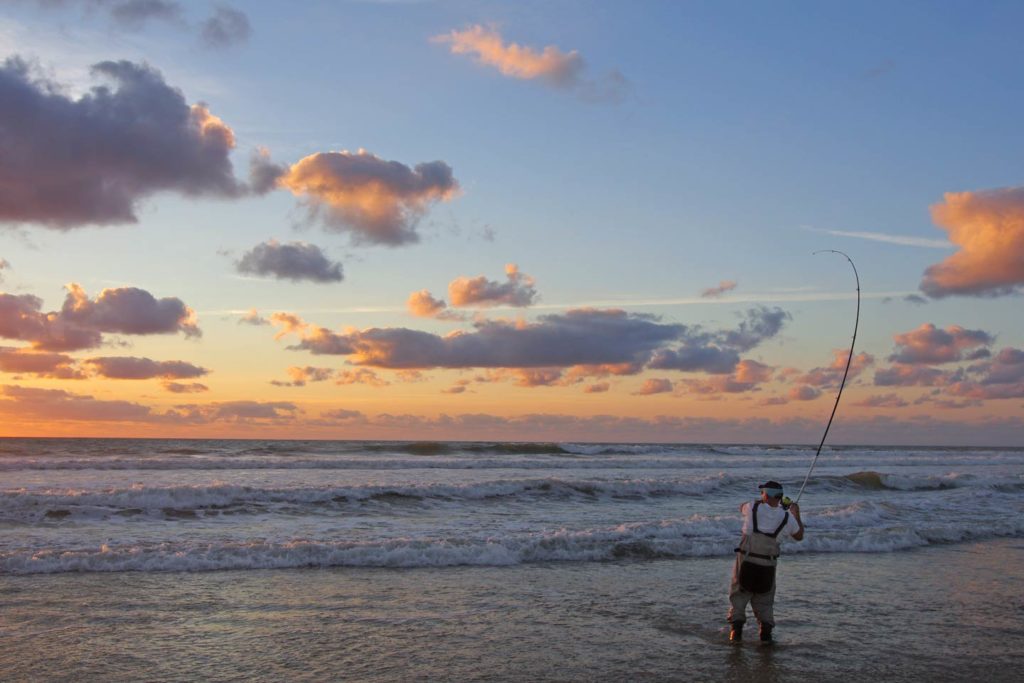 An angler casting into the surf at sunset