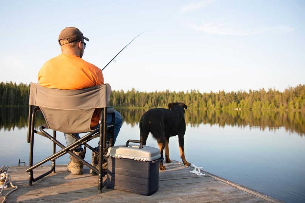 A view from behind of man sitting on a portable chair next to his dog, while casting a fishing rod into a calm body of water with some trees in the distance.