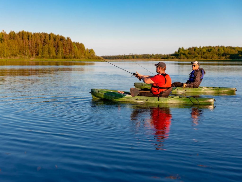 Two men in green kayaks, one casting a line, as he tries fishing in the Great Lakes at sunset, while the other looks on