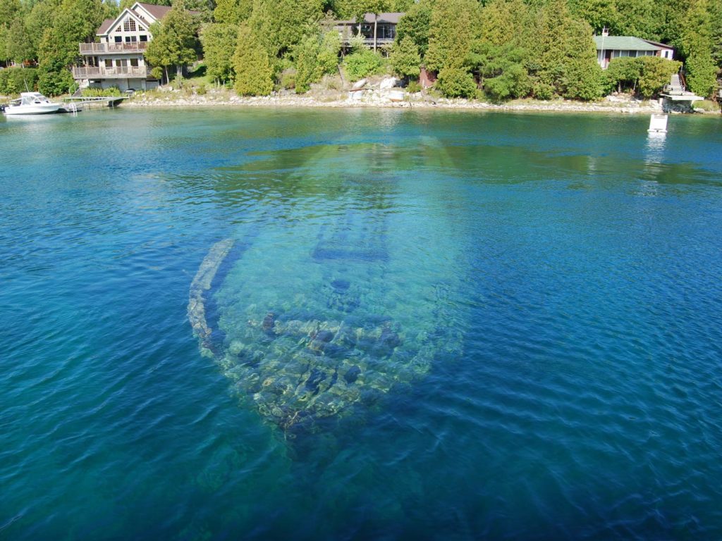 A view from above of a sunken ship in Lake Huron, visible beneath the crystal clear waters, with houses on the lake's shore visible in the background