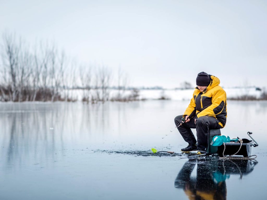 A man in a yellow coat, sitting on a box with another box full of fishing ear next to him, ice fishing in the Great Lakes on a grey day