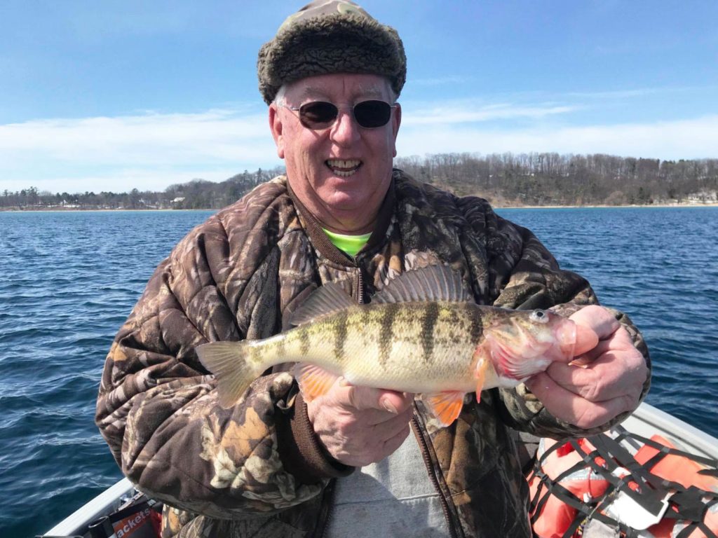 An angler in a furry hat, sunglasses, and a camouflage coat holding a Yellow Perch on a boat with the water and shoreline behind him