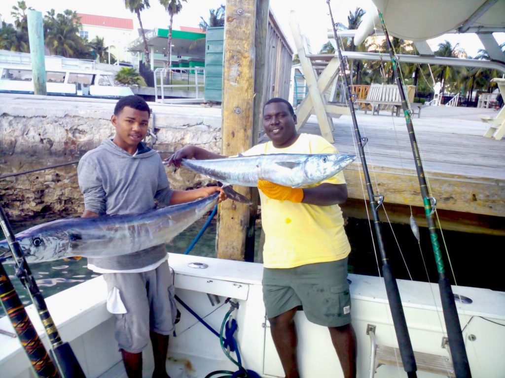 Two young fishermen standing on a fishing boat next to the dock, holding a large Wahoo each