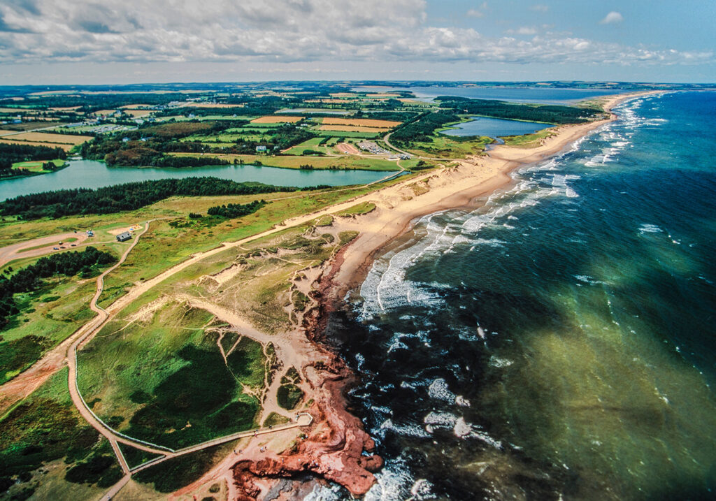 Eine Luftaufnahme der Prince Edward Island in Kanada mit gr&uuml;nen Feldern links, Strand in der Mitte und dunkelgr&uuml;nem Wasser rechts.