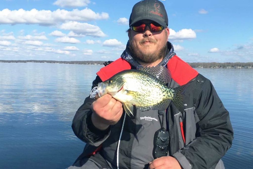 A man sits aboard a charter on a sunny day holding a Crappie to the camera