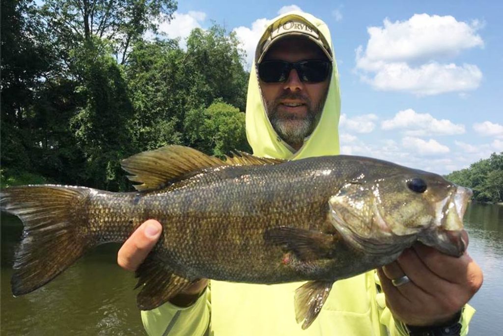 A man holds a Largemouth Bass up to the camera with greenery and the river behind him