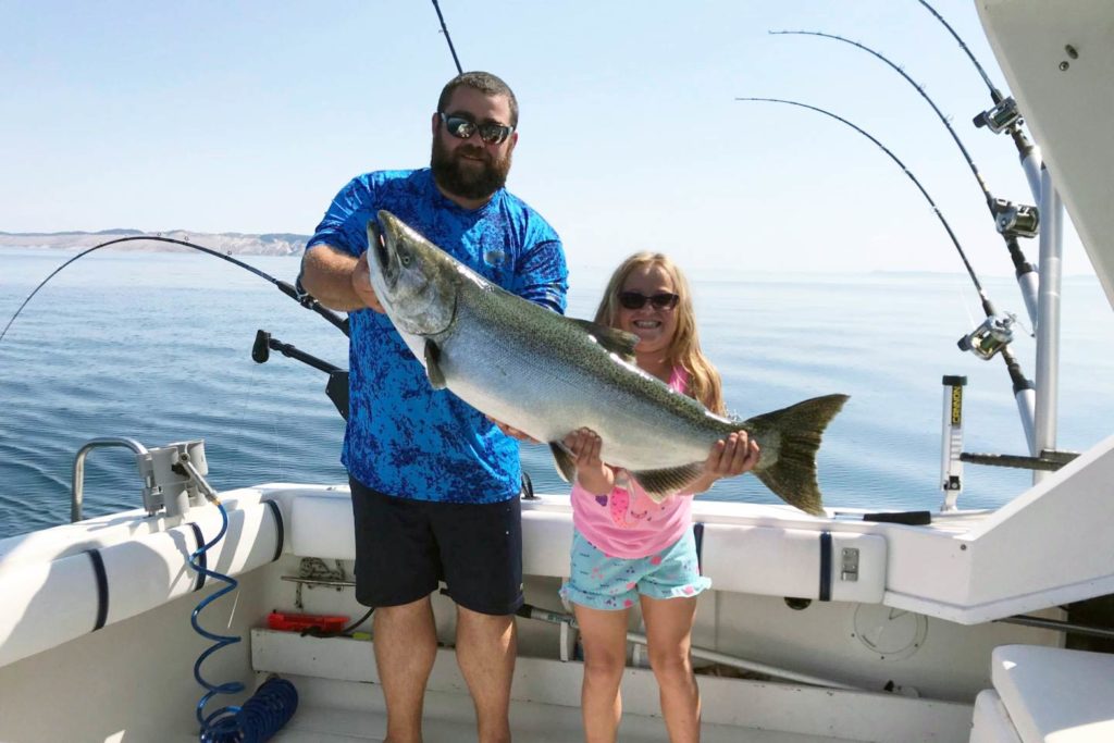 A man and a girl stand smiling on a charter boat with Lake Michigan behind them holding a large Chinook Salmon