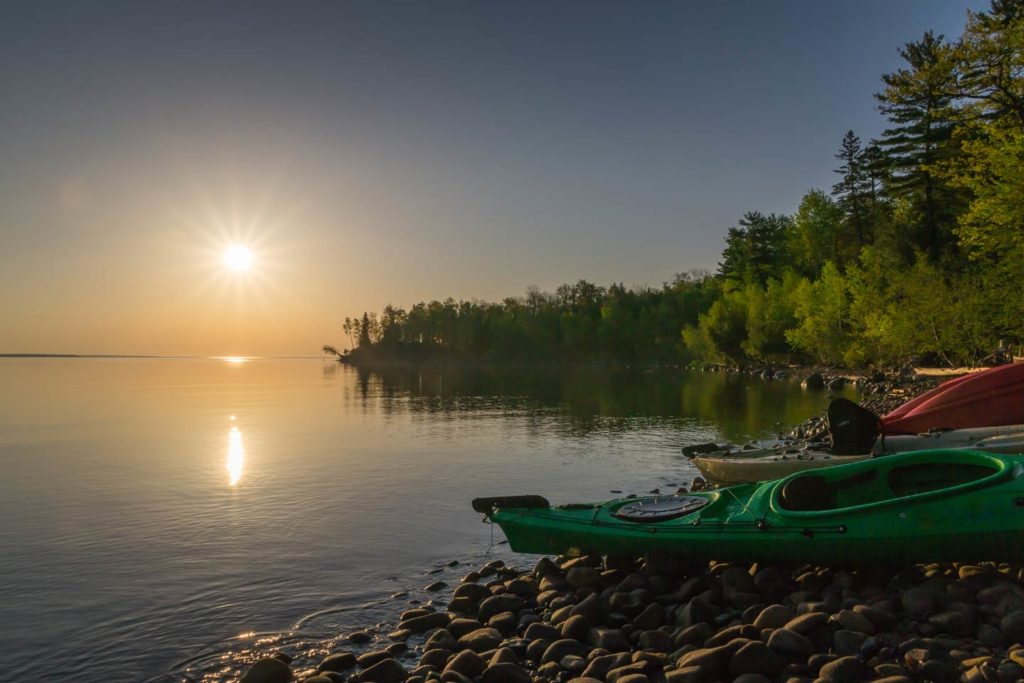 A view of a stone beach with kayaks placed on the ground and the lake in the background at sunset