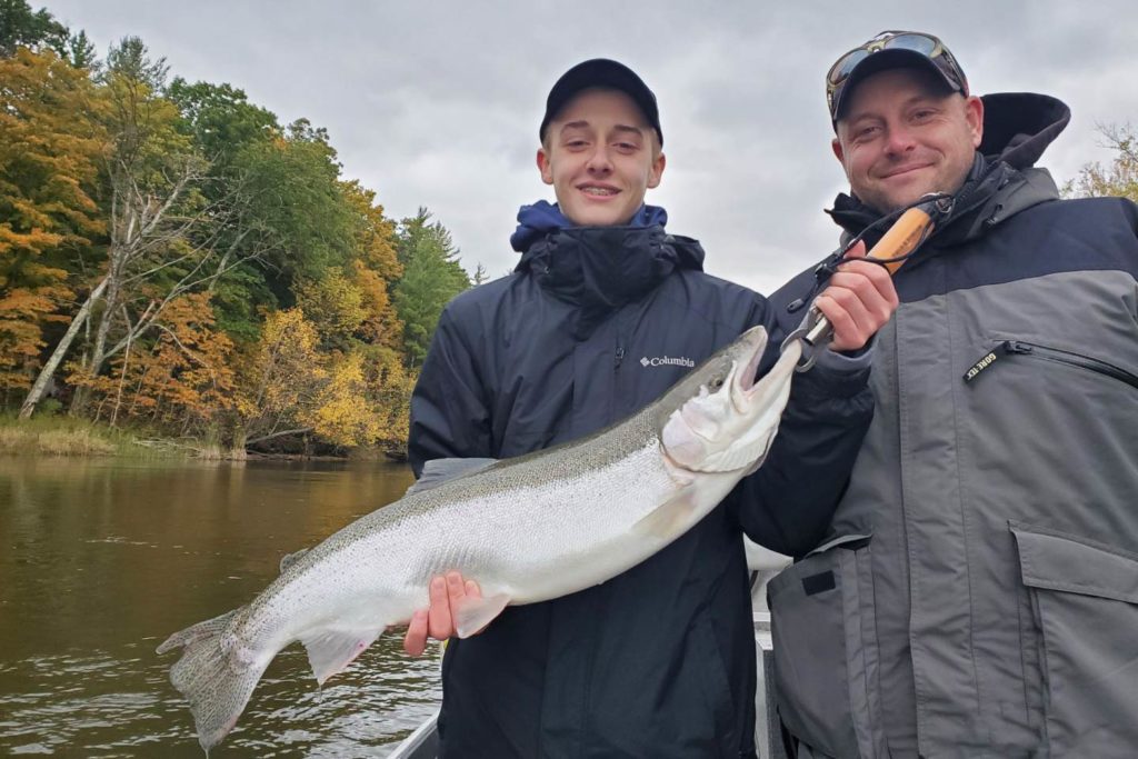 A man and a child hold a Steelhead up to the camera with the river behind them