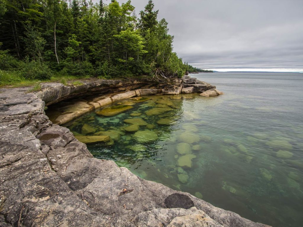 The shores of Lake Superior on a cloudy day, with clear waters and a rocky and green shoreline
