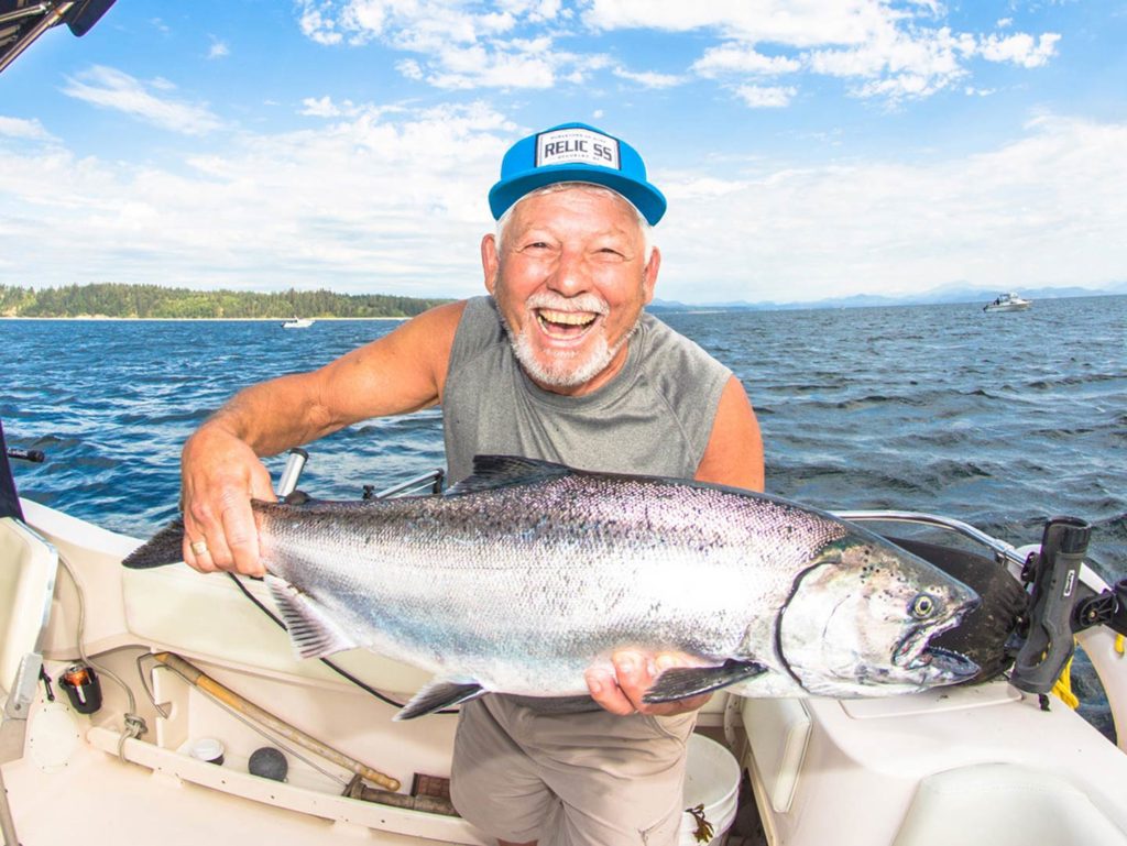 A happy elderly angler in a vest top holding a large Chinook Salmon on a boat, with the water and some land in the distance behind him
