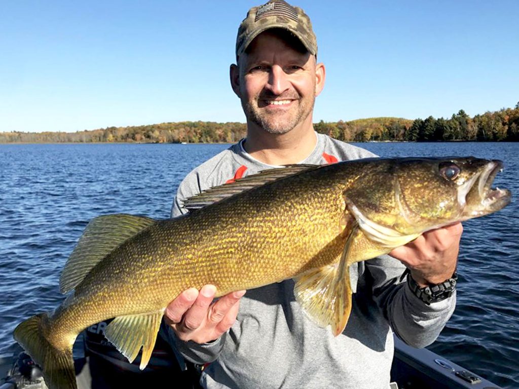 A happy angler on a boat in a khaki hat holding a Walleye caught fishing the Great Lakes