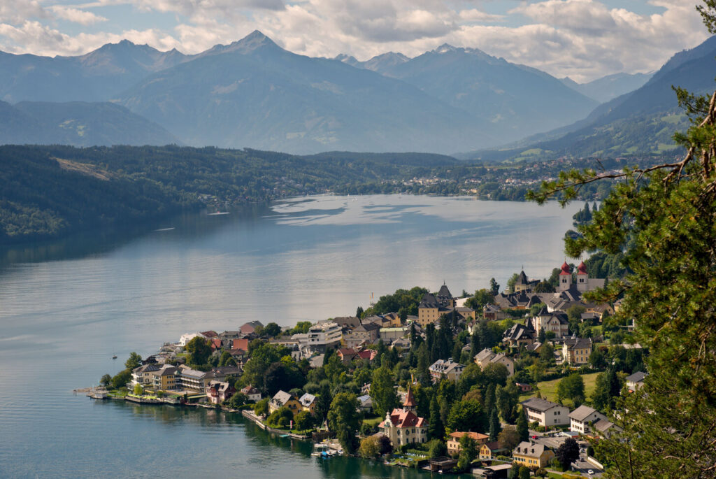 Ein Panorama des Millst&auml;tter Sees, mit den Alpen im Hintergrund und Millstatt auf einer Halbinsel auf der rechten Seite im Vordergrund.