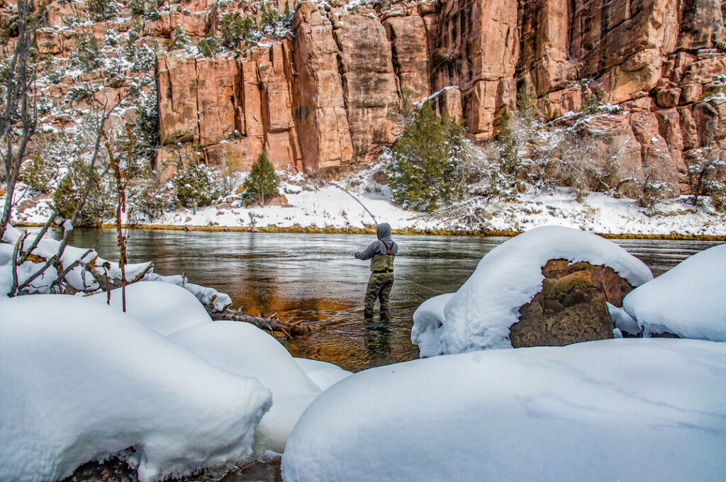 Ein Angler steht in einem Fluss zwischen schneebedeckten Felsen.