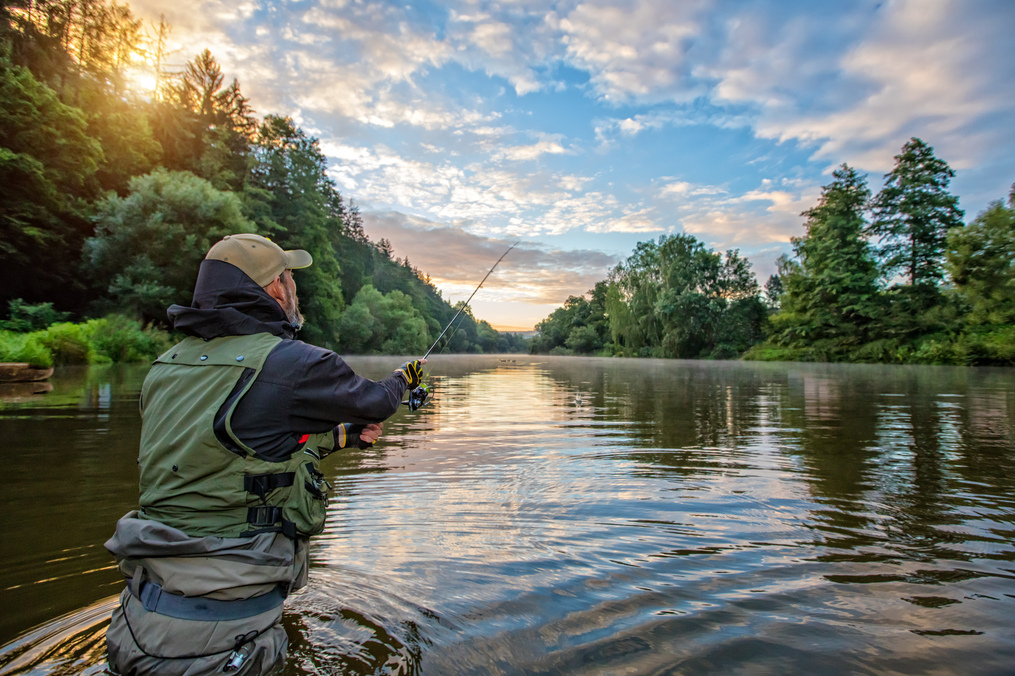 Ein Angler steht mit der Wathose in einem Gew&auml;sser und wirft seine Angel aus.
