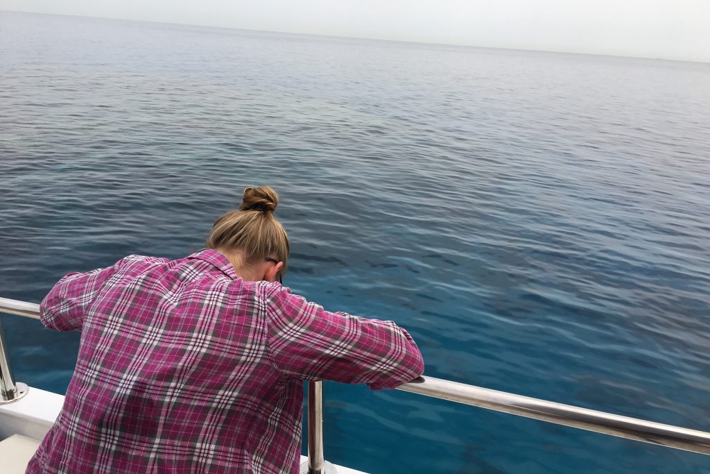 A person in a plaid shirt leans over the railing of a boat, looking down at the calm blue ocean water.