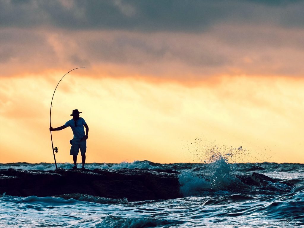 Angler steht bei Sonnenuntergang mit einer Angelrute in der Hand auf einem Steg.