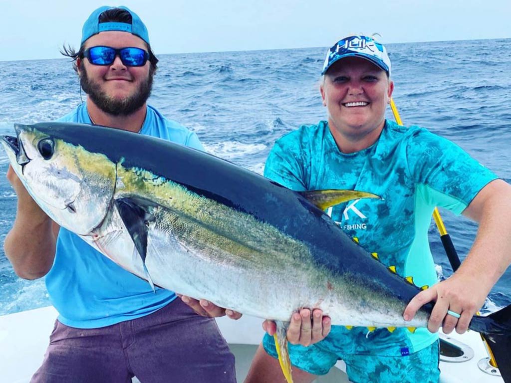 A photo of two smiling anglers sitting on a boat and holding a big Yellowfin Tuna