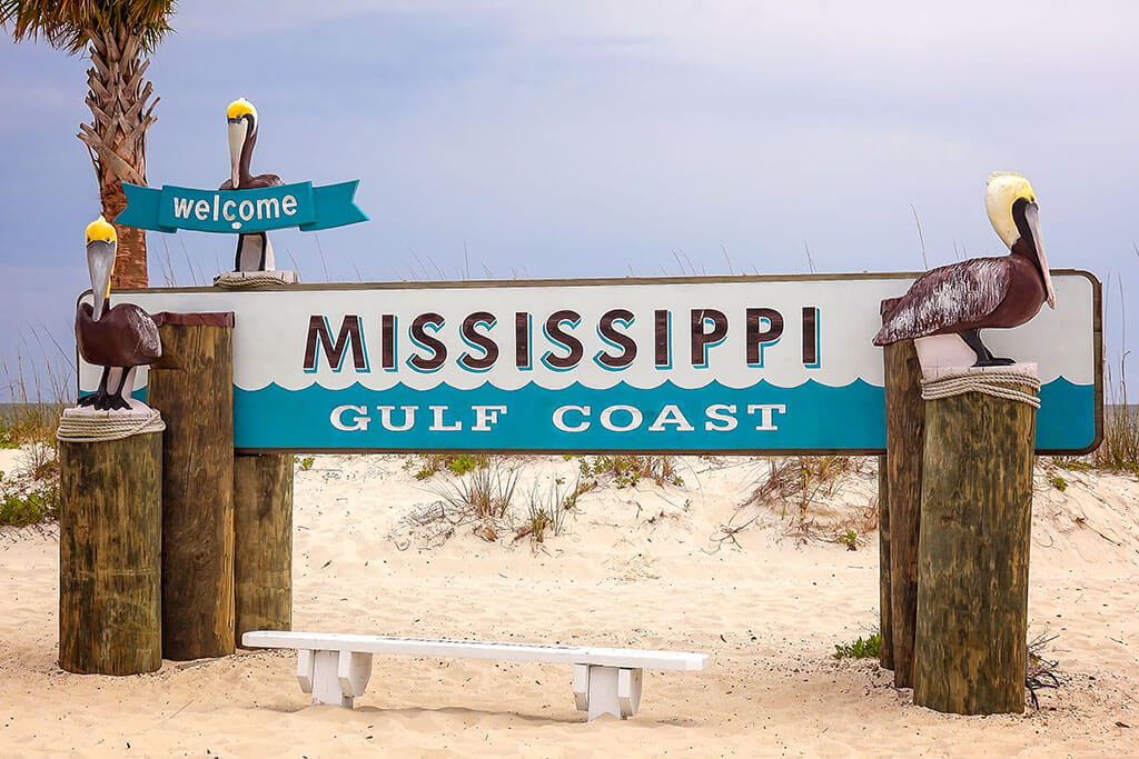 A wooden sign on a sandy beach that reads "Mississippi Gulf Coast", with a small white bench in front of it.