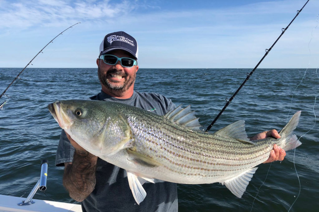 A man smiles as he holds a Striped Bass on a charter boat with the water behind him
