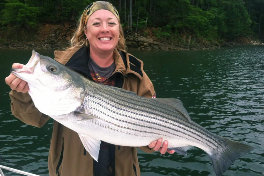 A woman smiles as she holds a Striped Bass on Lake Lanier with the lake behind her