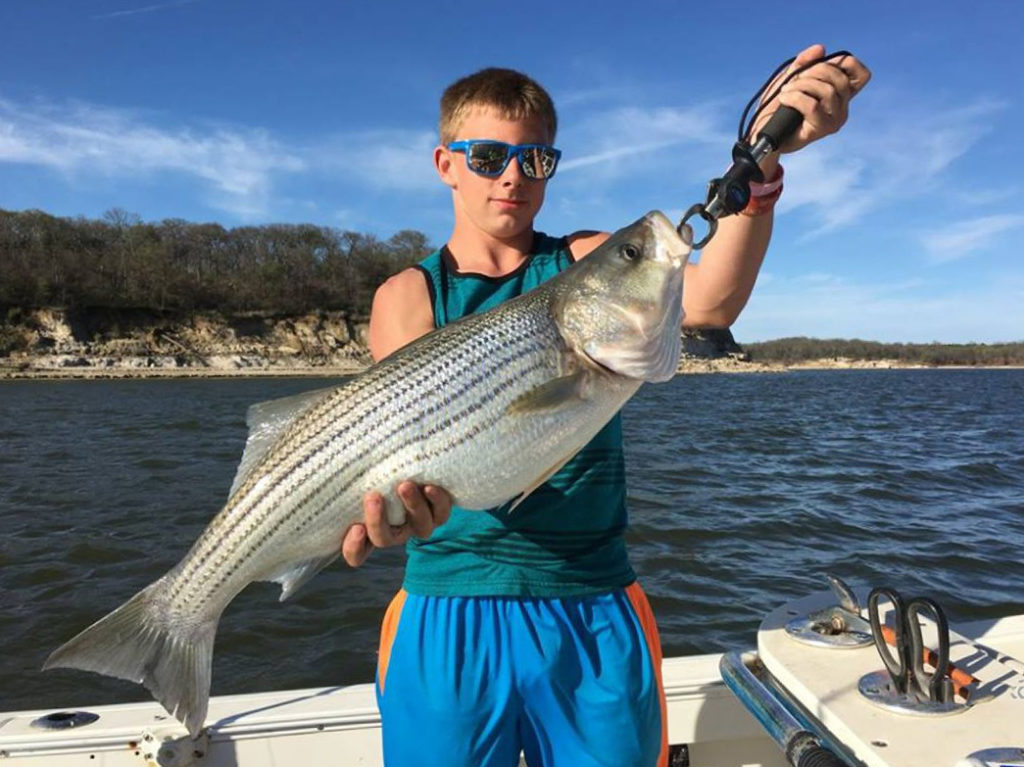 A teenage boy holds a large Striped Bass on a charter boat on a sunny day with water behind him