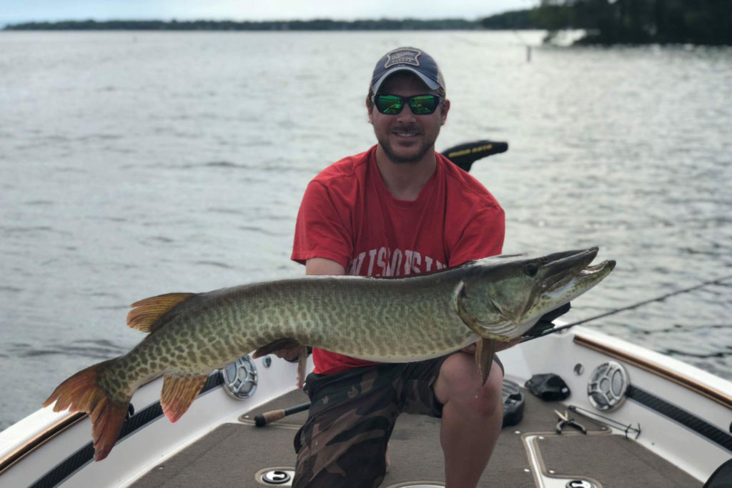 A man holds a Musky on board a charter boat with the lake behind him