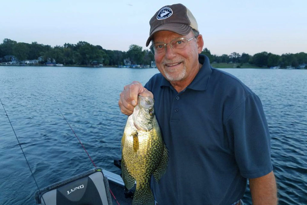 A man smiles and holds a Crappie on board a charter boat with the lake behind him
