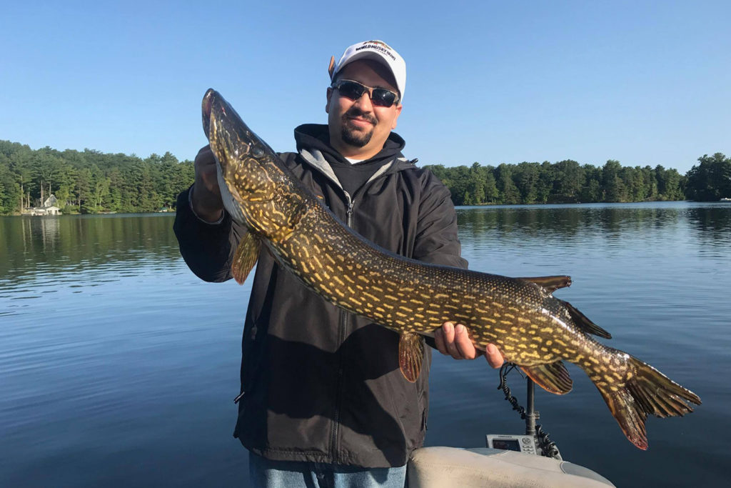A man holds a large Pike on a charter boat with the lake behind him