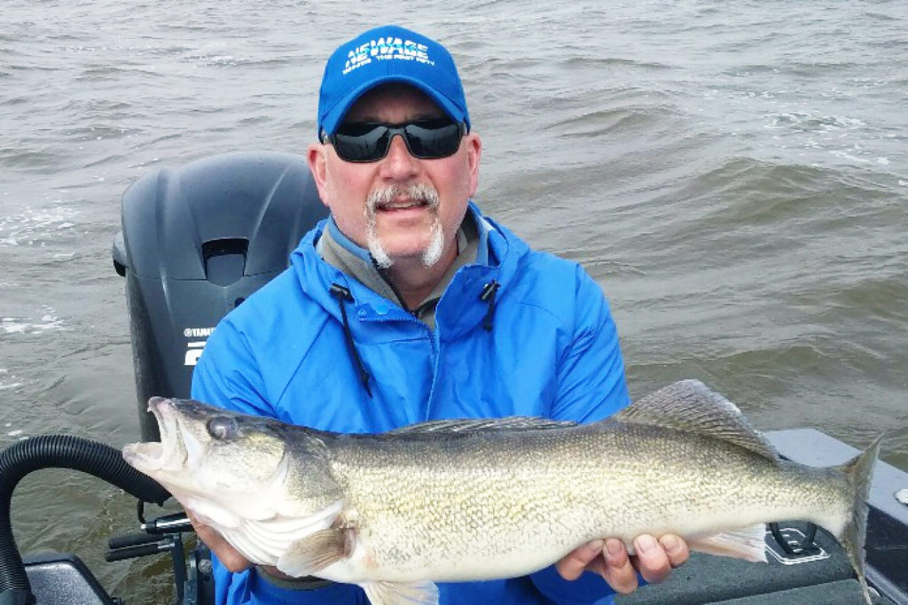 A man wearing sunglasses holds a large Walleye on board a charter vessel with the lake behind him