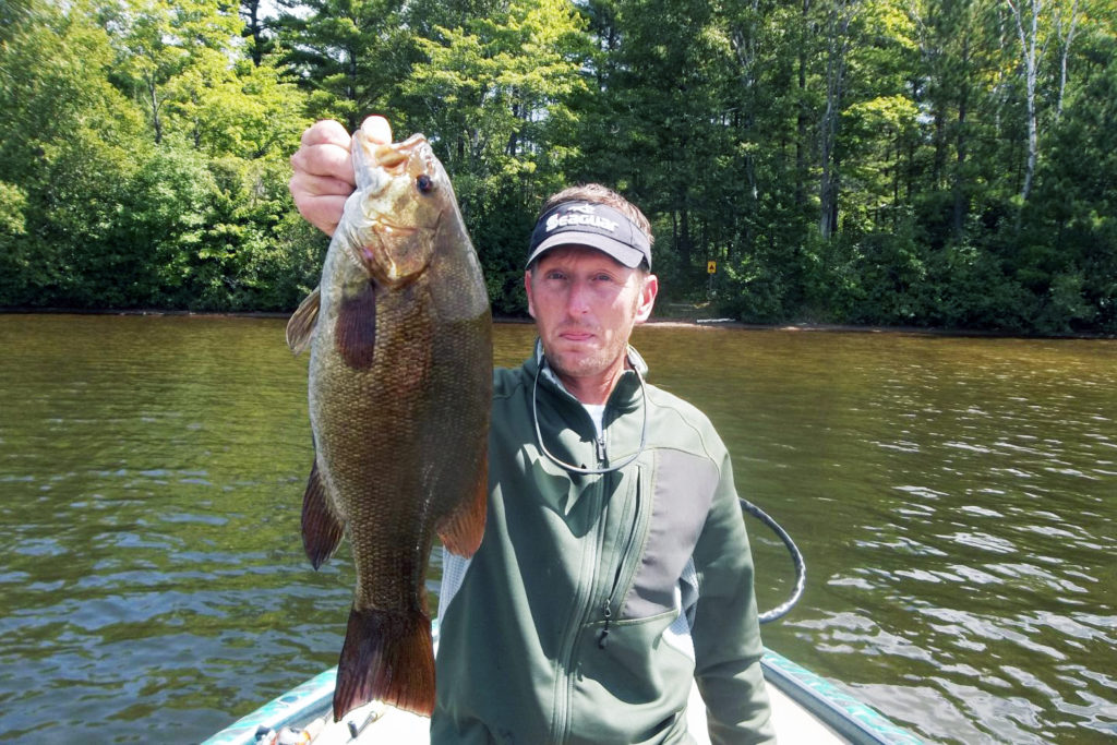A man holds a large Smallmouth Bass up to the camera with the lake behind him