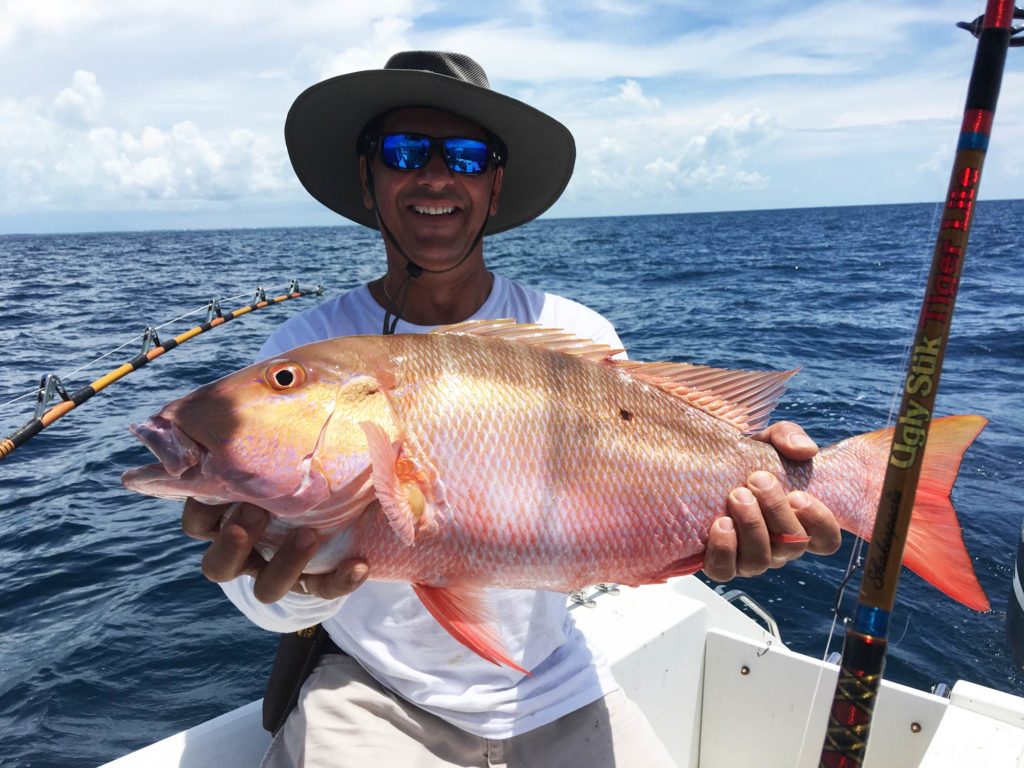 A smiling fisherman in a hat and sunglasses sitting on a boat, holding a big Mutton Snapper