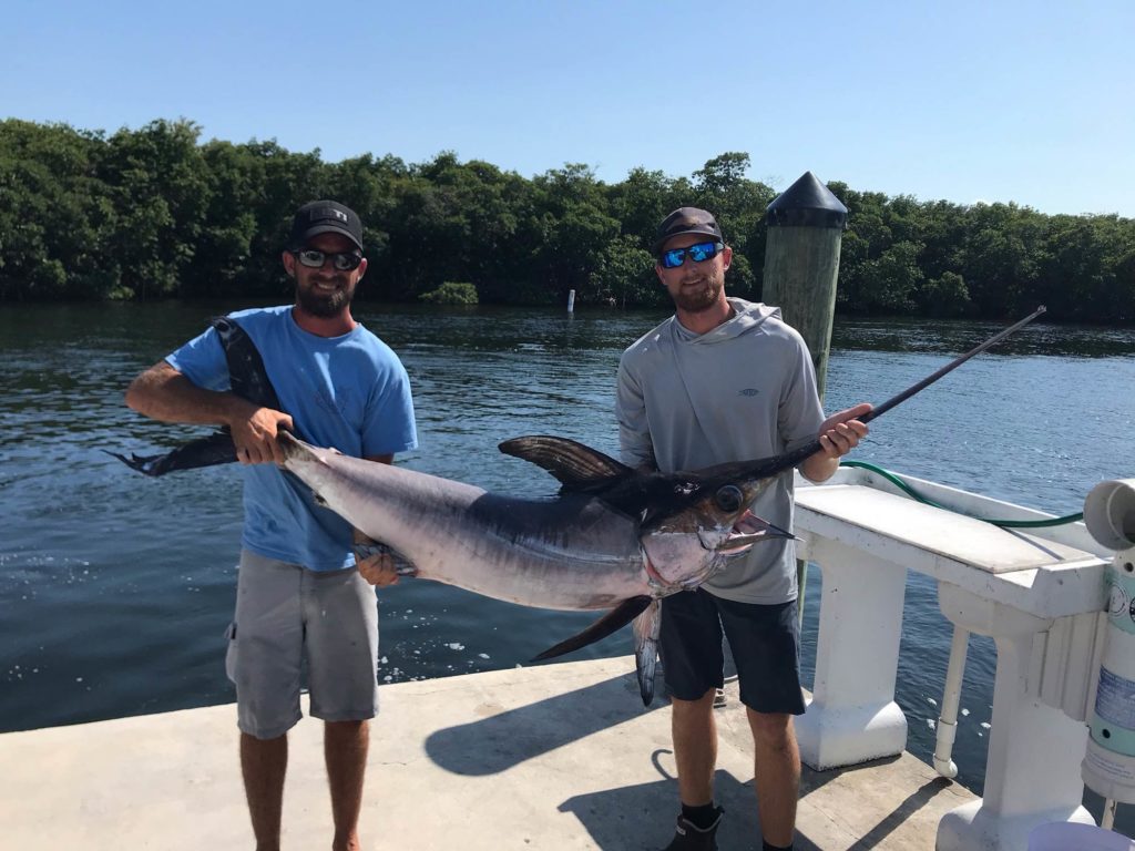 Two men standing on a dock, holding a Swordfish with water and mangroves in the background