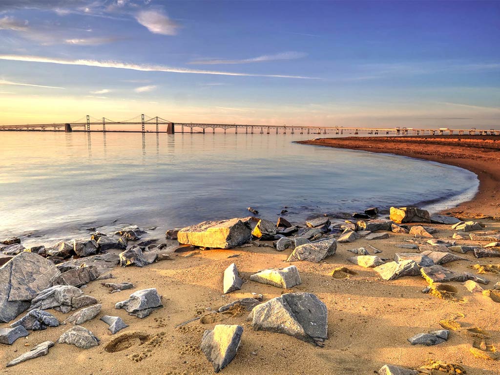 A coastal scene with a sandy beach and scattered rocks in the foreground captures the charm of Chesapeake Bay. In the background, a long bridge stretches across calm water under a clear sky, where there are just a few clouds. The sun casts a warm, golden light on the landscape.