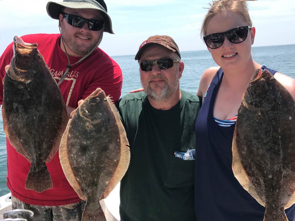 Three people on a boat proudly show off their Chesapeake Bay Flounder catches. The person on the left is wearing a red shirt and hat, the one in the middle has on a dark shirt and cap, and the one on the right is sporting sunglasses and a tank top. Clear skies stretch over the vast sea in the background.