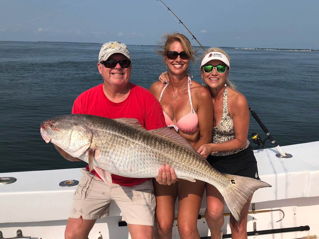 Three people are on a boat in Chesapeake Bay, proudly holding a large Redfish. The person on the left's wearing a red shirt and hat, the one in the middle's sporting a pink bikini, and the one on the right's wearing a patterned swimsuit with a white visor. Fishing rods suggest that more adventures await them on open water.