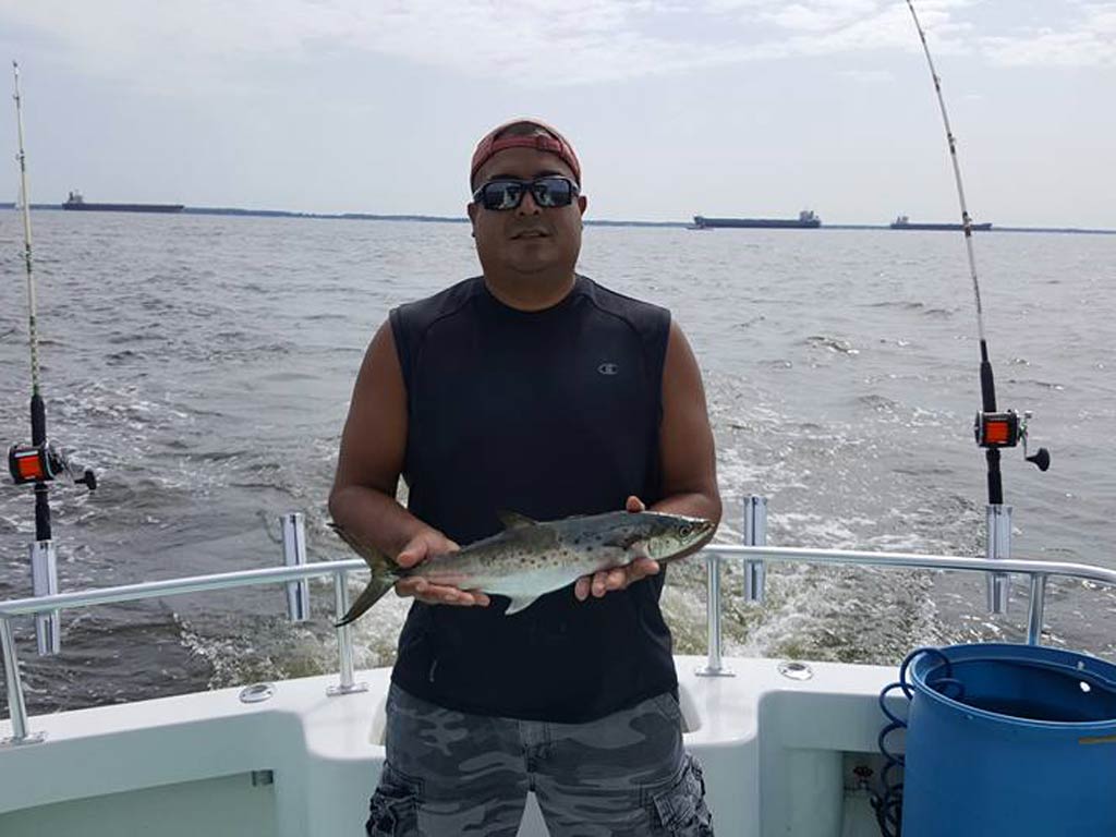 A person in a sleeveless shirt and a cap proudly holds Spanish Mackerel while standing on a boat. They’re enjoying Chesapeake Bay fishing, and other boats are visible in the distance. Fishing rods line the boat's railing, ready for action, while a blue container sits close by.
